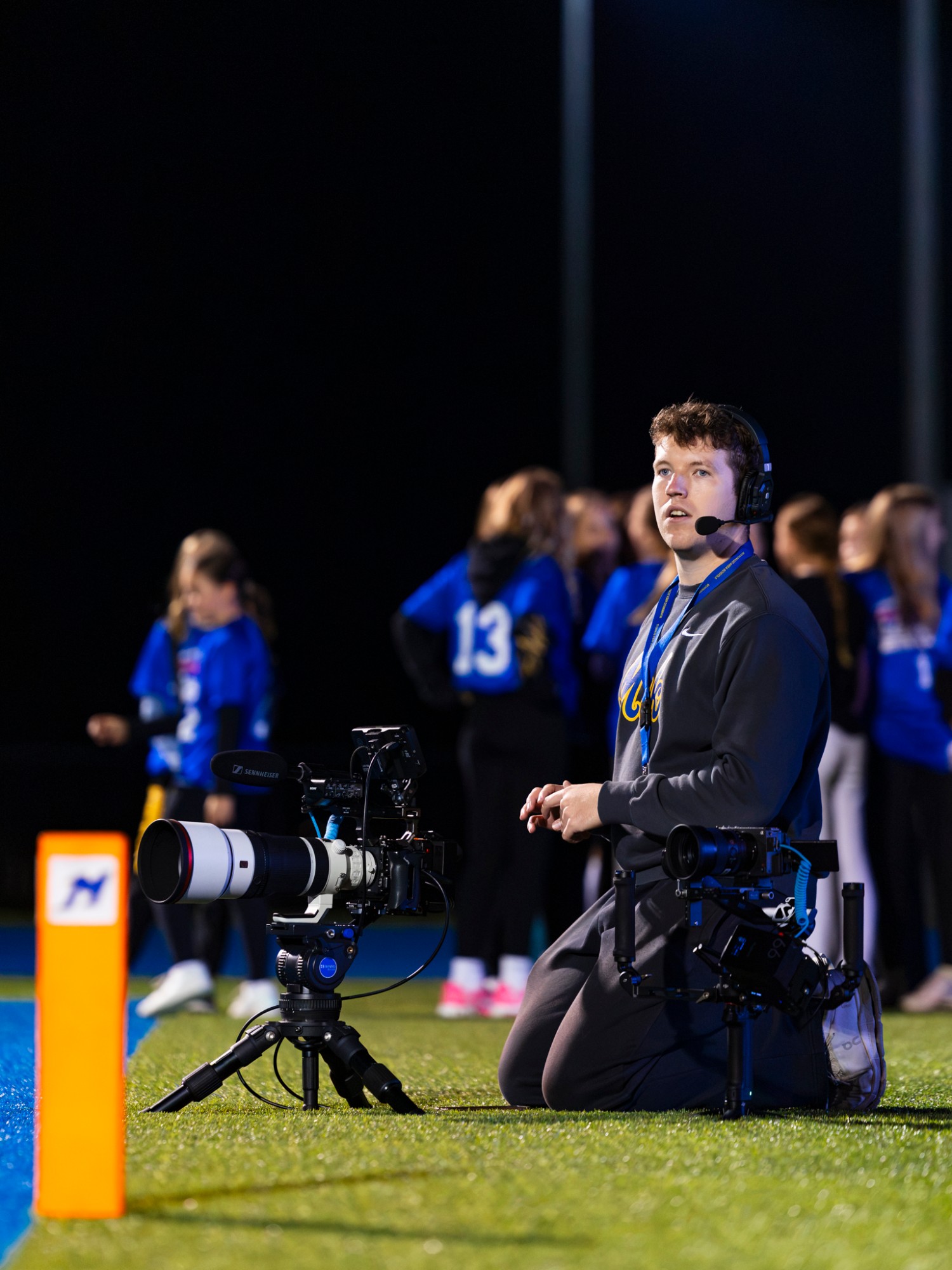 Ethan Jaworski kneeling on the sideline beside his cameras, wearing a headset